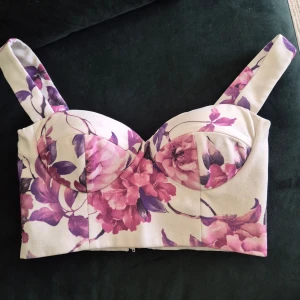 Blommig korsett-topp med breda axelband - Superfin korsett-topp med breda axelband och blommigt mönster i lila och rosa toner på vit botten. Toppen har formade kupor, dragkedja och hyskor i sidan för extra stöd. Perfekt för dig som vill sticka ut med en feminin och trendig look.