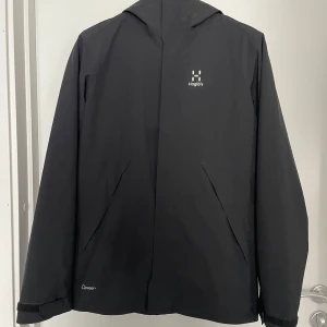 Svart vindjacka från Haglöfs - Svart vindjacka från Haglöfs med minimalistisk design och huva. Jackan har diskreta fickor framtill, dragkedja och justerbara ärmslut med kardborre. Materialet är syntet och jackan är perfekt för blåsiga dagar. Klassisk logga på bröstet och meshdetaljer på insidan.