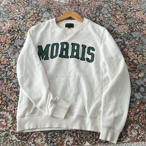 Vit sweatshirt från Morris med tryck - Vit sweatshirt från Morris med grönt tryck över bröstet. Klassisk rund halsringning, ribbade muddar och mjukt bomullsmaterial. Perfekt för en chill och stilren look. Snyggt tryck med MORRIS i grönt framtill.