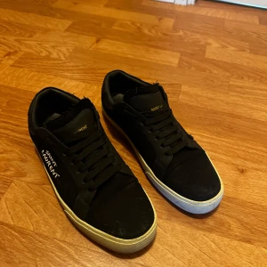 Svarta Saint Laurent sneakers - Svarta sneakers från Saint Laurent med vit logga broderad på sidan. Skorna har svarta snören, rund tå och vit platt sula. Ovandelen är i en mix av skinn och textil, vilket ger en clean och stilren look. Perfekta för dig som gillar exklusiva streetwear-vibbar.