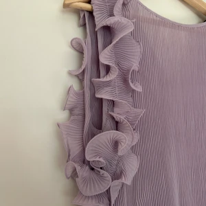 Ljuslila plisserad blus med volang - Superfin ljuslila blus med plisserat tyg och stora volangdetaljer längs ärmarna. Blusen har lång ärm och är tillverkad i ett lätt, transparent material som ger ett luftigt intryck. Perfekt för dig som vill sticka ut med en unik och trendig topp.