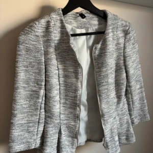 Gråmelerad kavajjacka med dragkedja - Snygg gråmelerad kavajjacka från H&M med dragkedja framtill. Jackan har en lätt figursydd passform, rundad halsringning och trekvartsärmar. Materialet är mjukt och strukturerat, perfekt för lager på lager. Passar dig som gillar stilrena och moderna plagg.