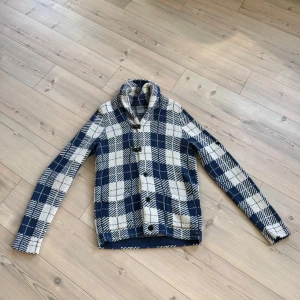 Blåvit rutig cardigan från J.C. Rags - Snygg cardigan från J.C. Rags i blåvitt rutigt stickat material. Jackan har en bred krage, coola spännen framtill och broderad logga på ärmen. Perfekt för dig som gillar street och vill sticka ut med mönster och detaljer.