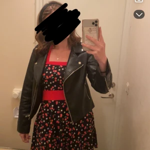 Svart kortklänning med körsbär från Akama - Säljer en svart kortklänning från Akama med rött midjeband och röda detaljer vid halsen. Klänningen har ett gulligt körsbärsmönster med vita stjärnor, är ärmlös och har en figurnära passform upptill med utställd kjol. Materialet är mjuk bomull, perfekt för sommarens alla tillfällen.