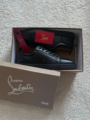 Svarta sneakers från Christian Louboutin - Säljer ett par svarta sneakers från Christian Louboutin i skinn med klassisk röd sula och diskret logga undertill. Skorna har rund tå, svarta snören och stilren design. Kommer med originalkartong och skohorn.