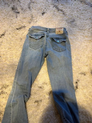 Ljusblå True Religion jeans bootcut 26 - Storlek 26 Säljer ett par ljusblå jeans från True Religion med ikoniska bakfickor och turkosa knappar. Klassisk bootcut-modell med normal passform och snygga detaljer på fickorna. Perfekta för dig som vill ha en avslappnad men ändå stilren look.