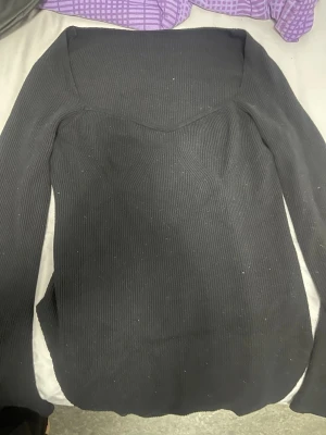 Svart ribbad långärmad topp - Säljer en svart ribbad topp med lång ärm och markerad sweetheart-ringning. Toppen är stretchig och har en figurnära passform som framhäver siluetten. Perfekt att styla med jeans eller kjol för en clean look.