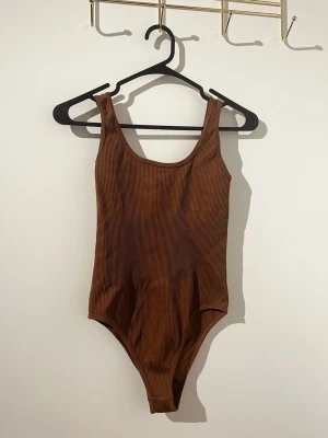 Brun ribbad shapewear body - Snygg oanvänd brun body med ribbad struktur och breda axelband. Modellen har en klassisk, rundad ringning och är tillverkad i ett stretchigt material som sitter skönt mot kroppen. Perfekt att styla med jeans eller kjol för en enkel men trendig look. 