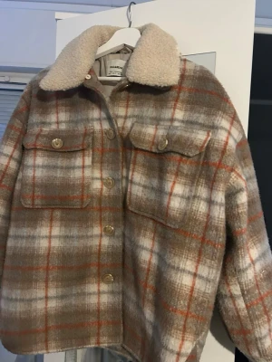 Rutig teddyjacka  - Säljer en mysig teddyjacka med rutigt mönster i beige, brunt och orange. Jackan har stora bröstfickor, knappar framtill och en fluffig krage i teddy. Perfekt för höst och vinter när du vill vara både varm och snygg.