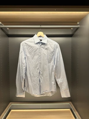 Ljusblå rutig skjorta från Riley - Snygg ljusblå skjorta från Riley i tailored fit med diskret rutigt mönster. Klassisk krage, långa ärmar och knappar framtill. Perfekt för dig som gillar stilrena och fräscha plagg.