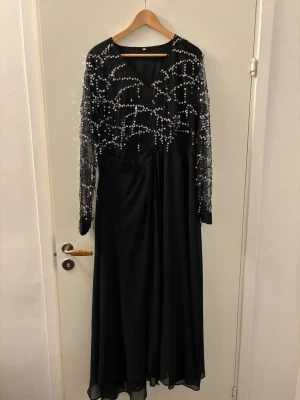 Svart långklänning med paljetter - Säljer en svart långklänning med transparenta långärmar och massor av glittriga paljetter på överdelen och ärmarna. Klänningen har v-ringning framtill och ett flytande, elegant fall i kjolen. Perfekt för dig som vill sticka ut med en glamorös look. det står 3XL men passar perfekt för L