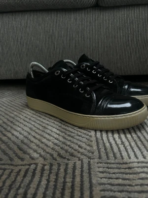Svarta Lanvin skor - Säljer ett par svarta Lanvin sneakers med ovandel i mocka och lackdetaljer på tån. Skorna har svarta snören, metallöljetter och en beige, platt gummisula. Klassisk och stilren modell som passar till mycket. Storlek 8