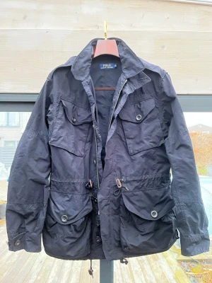Marinblå fieldjacket från Polo Ralph Lauren - Säljer en marinblå fieldjacket från Polo Ralph Lauren i storlek M sitter som L. Jackan har flera stora fickor med knappar, dragsko i midjan och dragkedja framtill. Tillverkad i ett lätt och slitstarkt syntetmaterial, perfekt för dig som vill ha en klassisk och funktionell jacka.