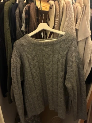 Grå stickad tröja från 157 - Mysig grå stickad tröja från 157 i storlek S. Tröjan har ett klassiskt flätmönster och är långärmad med rund halsringning. Perfekt för kyliga dagar och enkel att matcha med jeans eller kjol.