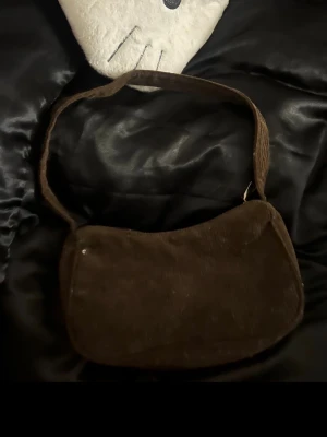 Brun manchesterväska med axelrem - Säljer en brun axelväska i manchester med bred axelrem och mjuk, rundad form. Väskan har ett enkelt, stilrent utseende och passar perfekt för dig som gillar retrovibbar och vill ha något unikt till din outfit.