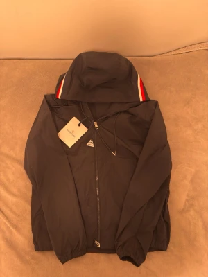 Svart Moncler vindjacka med huva - Säljer en svart vindjacka från Moncler med huva och dragkedja. Jackan har Moncler-logga på ärmen och snygga rödvita detaljer på huvan. Tillverkad i lätt polyester som passar perfekt när det blåser. Klassisk och stilren design med elastiska muddar. M MEN PASSAR S. PRIS KAN DISKUTERAS