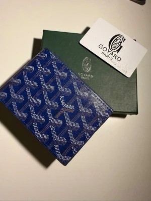 Blå Goyard plånbok med mönster - Säljer en blå plånbok från Goyard med deras ikoniska grafiska mönster i vitt och grått. Plånboken är kompakt och har en lyxig känsla med Goyard Paris-logga på framsidan. Perfekt för kort och sedlar, och levereras med originalask och äkthetskort.