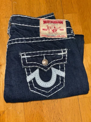 True Religion Billy Super T jeans blå - Äkta True Religion Billy Super T Row Seat jeans i mycket bra skick (nyskick). Klassisk modell med kraftiga Super T-sömmar och ljusblått broderi på bakfickorna. Tillverkade i USA i kvalitetsdenim med lätt stretch.  Detaljer: 	•	Storlek: W31 M/L 	•	Material: 99 % bomull, 1 % spandex 	•	Färg: Mörkblå 	•	Made in USA  Mått (platt mätt): 	•	Midja: 40 cm 	•	Innerben: 82 cm 	•	Ytterlängd fram/bak: 102 / 107 cm
