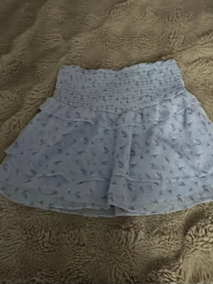 Ljusblå blommig volangkjol - Supersöt ljusblå kortkjol med små blommor och volangdetaljer. Kjolen har en bred, smockad midja som ger en bekväm passform och ett flowy fall. Perfekt för varma dagar och ger en romantisk vibe till din outfit.