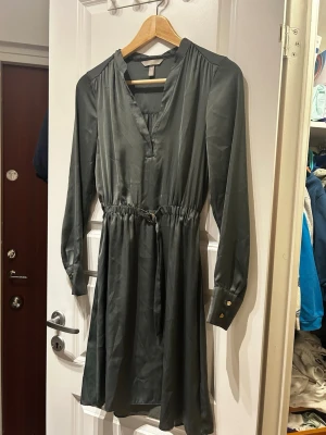 Mörkgrön satin-klänning från H&M - Supersnygg mörkgrön klänning i satin från H&M med v-ringning, långa ärmar och markerad midja med knytband. Klänningen har guldiga knappar vid ärmsluten och ett lätt glansigt material som ger en lyxig känsla.