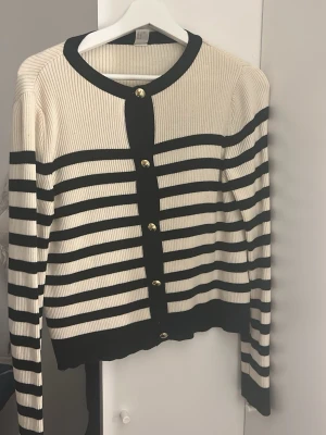 Randig kofta med guldknappar - Snygg ribbad kofta i beige och svart med horisontella ränder och dekorativa guldknappar framtill. Långärmad och med svart kant vid hals, knappslå och nederkant. Perfekt för lager-på-lager och ger en stilren look.