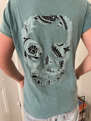 Grön t-shirt med döskalle och knappar - Säljer en grön t-shirt i mjuk bomull med korta ärmar. På ryggen finns en stor döskalle med blommigt mönster i svart och ljusblått. Framtill har tröjan en v-ringning med små knappar längs kanten. Skön och avslappnad passform.