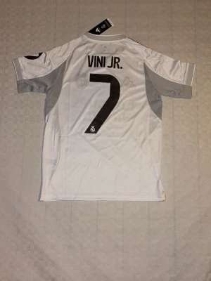 VINICIUS JR REAL MADRID TRÖJA. -  En vit Real Madrid matchtröja med Vini Jr. och nummer 7. Tröjan har svarta och gula detaljer samt Adidas-logga.