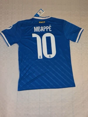 Blå Real Madrid Mbappé tröja Adidas - Säljer en blå Real Madrid matchtröja med Mbappé och nummer 10 på ryggen. Tröjan har vita detaljer, klubbmärke, Adidas-logga och sponsortryck framtill. Tillverkad i polyester med klassiska ränder på axlarna och vit krage.
