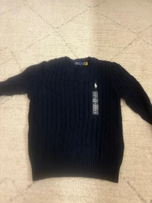 Mörkblå stickad tröja Polo Ralph Lauren - Säljer en mörkblå kabelstickad tröja från Polo Ralph Lauren med klassisk rund hals och broderad logga på bröstet. Tröjan har lång ärm och är aldrig använd och i väldigt bra skick!