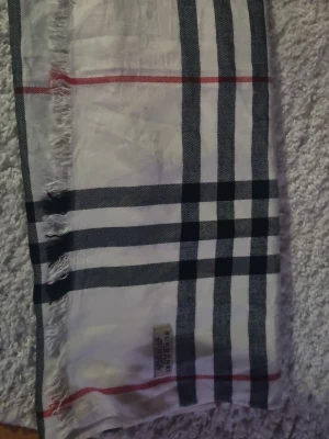 BBurberry kashmir halsduk, äkta och använd 1 gång - Snygg väst från Burberry i klassiskt rutigt mönster med vita, svarta och röda toner. Västen är tillverkad i 100% kashmir och har fransade kanter. Perfekt för dig som vill ha en tidlös och lyxig look.