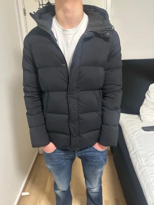 Svart pufferjacka med huva - Säljer en svart J. Linderberg jacka med huva och dragkedja framtill. Jackan är vadderad och har en stilren look med raka linjer och diskreta fickor. Perfekt för kalla dagar och enkel att matcha med olika outfits.