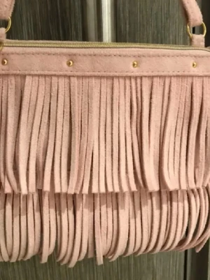 Rosa axelväska med fransar i mocka - Säljer en superfin rosa axelväska i mocka med långa fransar och guldfärgade detaljer. Väskan har dragkedja upptill och en justerbar axelrem. Perfekt för dig som vill sticka ut med en cool accessoar!