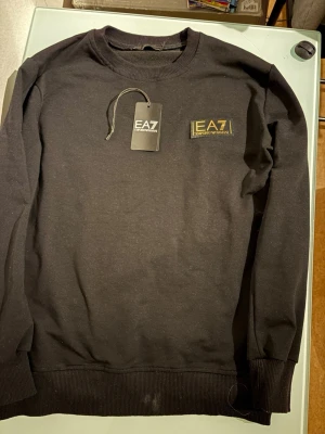 Svart EA7 Emporio Armani tröja - Svart crewneck-tröja från EA7 Emporio Armani med guldfärgad logga på bröstet. Tröjan har ribbade muddar vid ärmslut och nederkant, klassisk rund hals och är tillverkad i mjuk bomull. Perfekt för dig som gillar stilrena och exklusiva plagg.