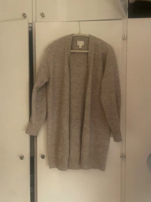 Beige lång kofta från H&M - Mysig och lång beige kofta från H&M med öppen front och ribbade muddar. Perfekt att slänga på sig över en t-shirt eller hoodie när det är kyligt. Koftan är stickad och har en avslappnad passform med långa ärmar.