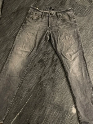Grå  jeans från Jack & Jones - Säljer ett par grå slim jeans från Jack & Jones, modell Glenn. De har klassisk femficksdesign, bälteshällor och knappgylf. Jeansen är i stretchigt material och har en snygg tvättad look med ljusa partier.