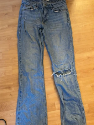 Blå straight jeans från Perfect Jeans - Säljer ett par klassiska blå jeans från Perfect Jeans i rak modell. Jeansen har slitningar och ett stort hål på ena benet för en riktigt edgy look. De har fyra fickor, normal passform och är tillverkade i robust denim. Perfekta för dig som gillar vintagevibbar och streetstyle.