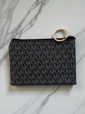 Svart plånbok från Michael Kors - Snygg svart plånbok från Michael Kors med klassiskt MK-mönster över hela ytan. Plånboken har dragkedja upptill och en guldfärgad ring som detalj. Perfekt storlek för kort och smågrejer, och riktigt stilren design.