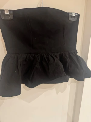 Svart volang tubtopp - Snygg svart tubtopp med volang nertill. Toppen är axelbandslös och har en bandeauformad överdel som sitter tight mot kroppen. Tillverkad i ett mjukt bomullstyg som känns bekvämt mot huden. Perfekt att styla med jeans eller kjol för en trendig look.