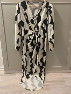 Svartvit långklänning med vågmönster - Säljer en svartvit långklänning med coolt vågigt mönster. Klänningen har omlott-skärning, v-ringning och långa ärmar med resår vid handlederna. Materialet är glansigt och känns som polyester. Perfekt för dig som vill sticka ut!