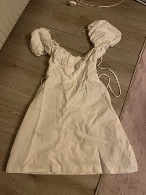 Vit kort klänning med puffärmar - Supersöt vit kort klänning med puffiga ärmar och snörning i ryggen. Klänningen har en feminin passform och är gjord i ett lätt bomullstyg, perfekt för varma dagar. Snygg v-ringning och fina detaljer som ger en romantisk vibe. Jätte fin som avslutningsklänning eller studentklänning.