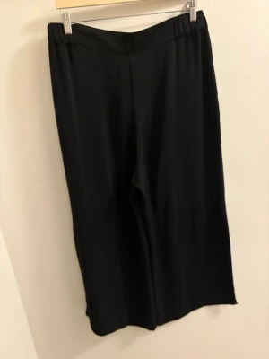 Svarta vida kostymbyxor - Säljer ett par svarta kostymbyxor med breda ben och hög midja. Byxorna har en klassisk look med rak passform och är tillverkade i ett mjukt, lätt material som ger en snygg drapering. Perfekta till en stilren outfit.