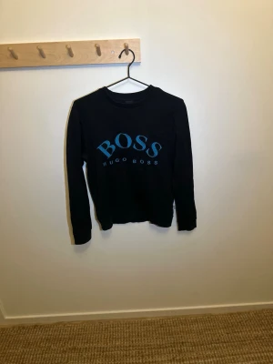 Svart sweatshirt från Hugo Boss - Säljer en svart sweatshirt från Hugo Boss med stor blå broderad logga på bröstet. Tröjan har rund halsringning och långa ärmar. Tillverkad i mjuk bomull för skön känsla och stilren look.  Skick 10/10!!