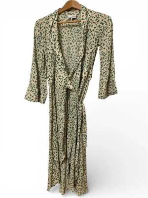 Beige blommig omlottklänning Ganni - Säljer en lång beige omlottklänning från Ganni med grönt blommönster. Klänningen har trekvartsärm, knytband i midjan och en avslappnad passform. Perfekt för dig som gillar en feminin och trendig stil.