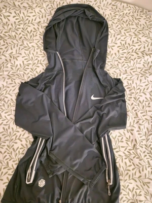 Gråish blå Nike vindjacka med huva - Säljer en gråish blå vindjacka från Nike med huva och dragkedja framtill. Jackan har vita detaljer, två fickor med dragkedja och är tillverkad i lätt polyester. Perfekt för utomhusaktiviteter och träning. Ny skick, använda kanske 2-3 gånger. Junior storlek M men passar som XS