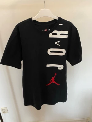 Svart Jordan t-shirt med tryck, storlek Medium  - Säljer en svart t-shirt från Jordan med stort vitt JORDAN-tryck längs framsidan och baksidan samt den klassiska röda Jumpman-loggan. T-shirten har rund hals, korta ärmar och är gjord i 100% bomull för skön känsla.