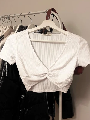 Vit ribbad croppad topp Pull&Bear - Säljer en vit croppad topp från Pull&Bear i ribbad bomullsmix. Toppen har korta ärmar, v-ringning och en snygg twistad detalj framtill. Perfekt till jeans eller kjol för en fräsch och trendig look.