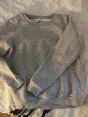Grå sweatshirt från Stories - En basic grå sweatshirt från Stories med rund hals och långa ärmar. Tröjan har ribbade muddar vid ärmslut och nederkant, perfekt för en chill och avslappnad stil. Tillverkad i mjuk bomullsmix som är skön att ha på sig.