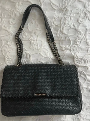 Svart axelväska från Valentino - Snygg svart axelväska från Valentino med flätad design och kedjedetaljer på axelremmen. Väskan är tillverkad i skinn och har flera fack inuti, inklusive ett med dragkedja. Perfekt för dig som vill ha en stilren och trendig väska med plats för det viktigaste. OBS! Se defekt på bild 5!!