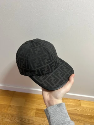 Fendi Keps - Säljer en svart keps från Fendi med det klassiska FF-logomönstret över hela kepsen. Kepsen har böjd skärm och justerbar rem baktill med Fendi Roma-detalj. Materialet är bomull och polyester, vilket gör den både snygg och bekväm.