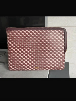 Mönstrad datorväska i vinrött - Snygg datorväska i vinröd ton med vitt geometriskt mönster. Väskan har dragkedja upptill och är tillverkad i ett tåligt syntetmaterial. Perfekt för att bära din laptop med stil.